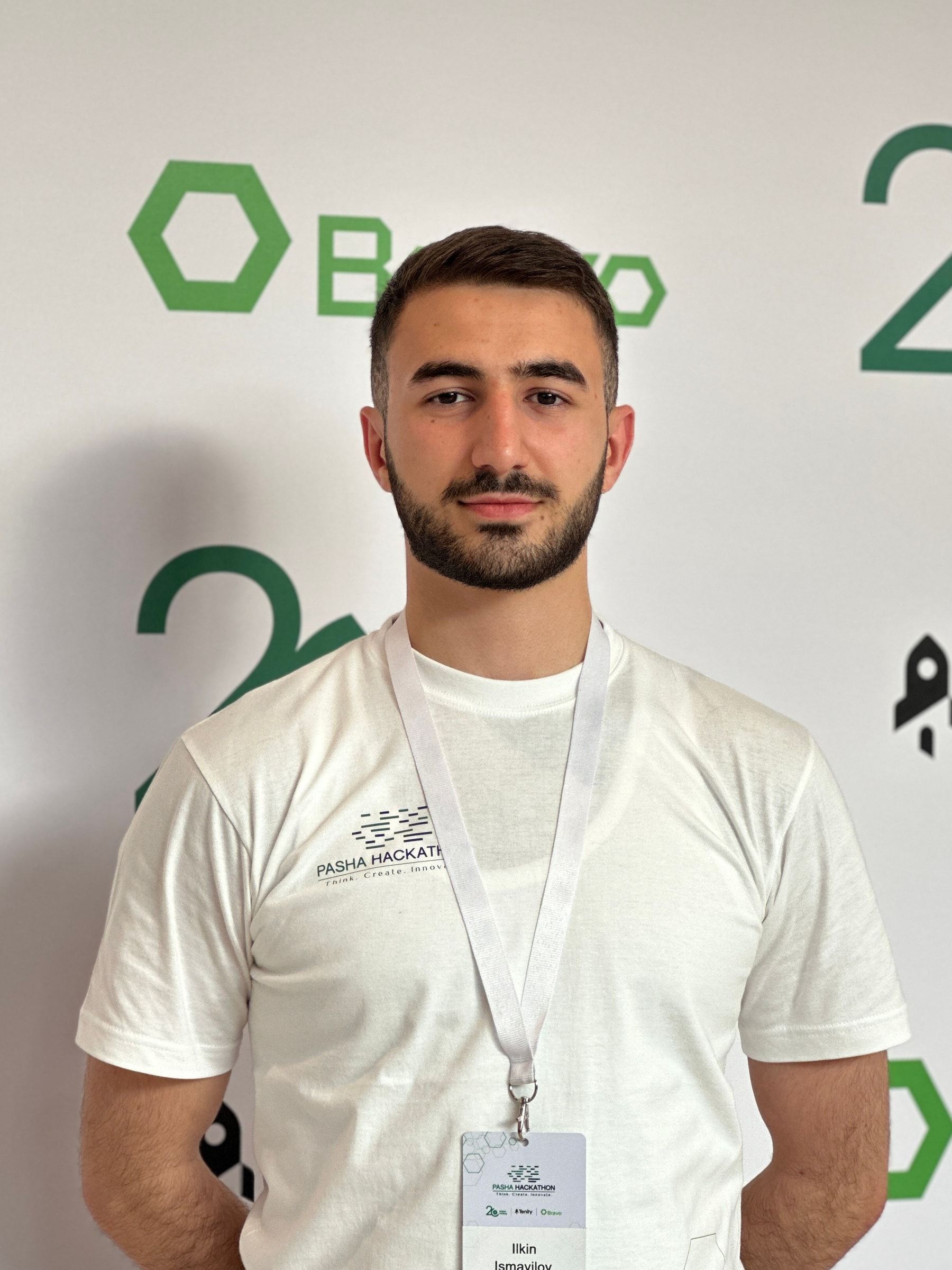 Ilkin Ismayilov in a white PASHA Hackathon T-shirt with a name badge, standing in front of a sponsor wall.